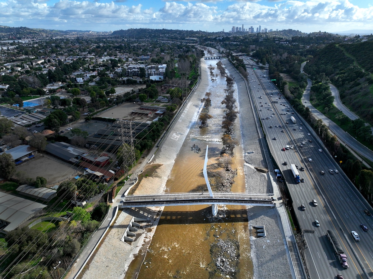 Aerial photo of water flowing down the Los Angeles River amid a break in heavy atmospheric rain with a view of the North Atwater Bridge linking Atwater Village with Griffith Park, with a view of the downtown Los Angeles skyline in the distance